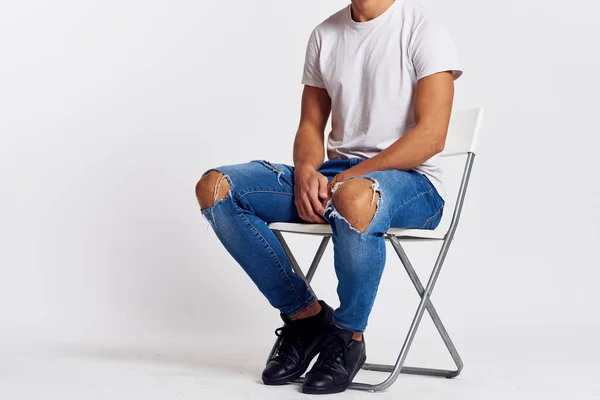 Un uomo in jeans e una t-shirt si siede su una sedia su uno sfondo leggero vista laterale — Foto Stock