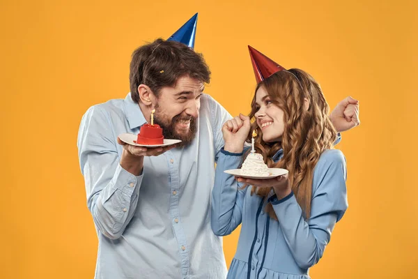 Homem e mulher em uma festa em bonés e com bolo de ouropel divertido fundo amarelo — Fotografia de Stock