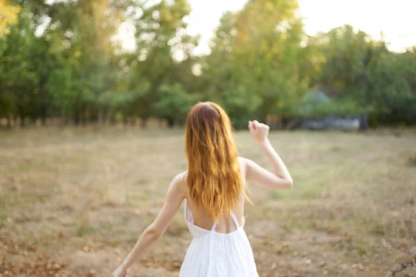 Beyaz elbiseli mutlu kızıl saçlı kadın ağaçların yanındaki kuru çimlerde koşuyor. — Stok fotoğraf