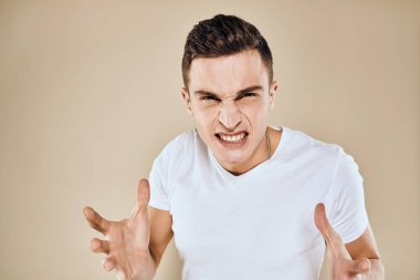 Yüzünde hoşnutsuzluk ifadesi olan beyaz tişört jestleri ve bej arka planı olan bir adam.