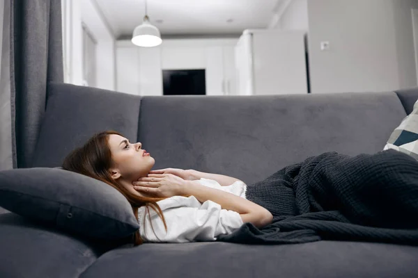 Mujer sosteniendo su cuello mientras está acostada en el sofá escondida con una manta en casa sintiéndose mal —  Fotos de Stock
