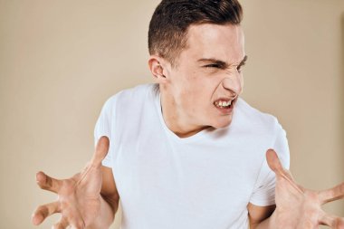 Yüzünde hoşnutsuzluk ifadesi olan beyaz tişört jestleri ve bej arka planı olan bir adam.