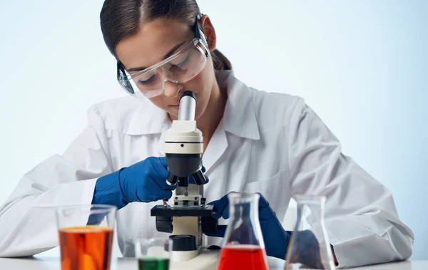 Assistente de laboratório feminino com um microscópio na mesa e óculos no líquido do rosto em um frasco — Fotografia de Stock
