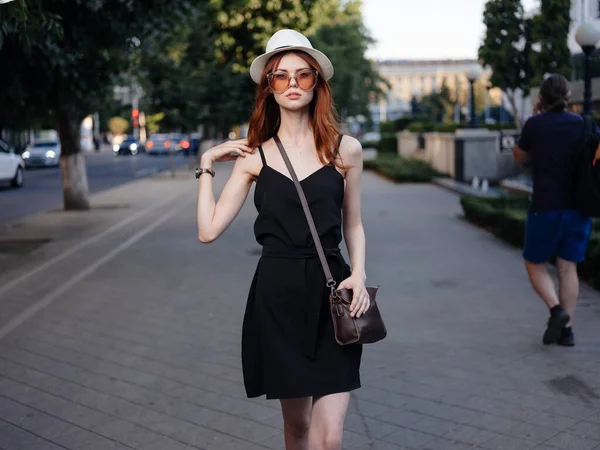 Una mujer con un vestido negro y un sombrero blanco con una bolsa en el hombro en la calle — Foto de Stock