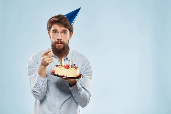 Uomo barbuto con un piatto di torta su sfondo blu e un cappello da festa di compleanno sulla testa — Foto Stock