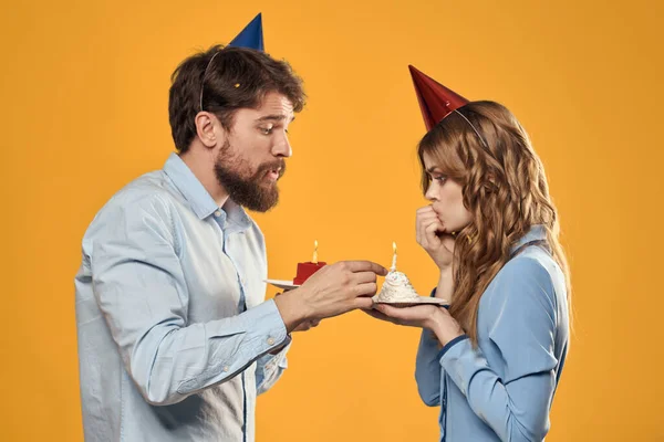 Γεννέθλια κόμμα άνθρωπος και γυναίκα σε ένα κίτρινο φόντο σε καπέλα με ένα κέικ στα χέρια τους — Φωτογραφία Αρχείου