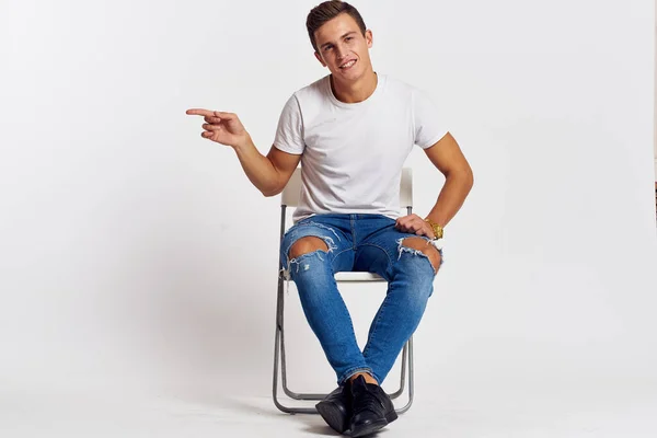 Mann auf einem Stuhl drinnen zerrissene Jeans weißes T-Shirt hübsches Gesicht Modell hellen Hintergrund — Stockfoto
