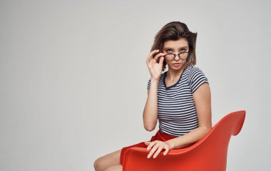 Etekli ve çizgili tişörtlü bir kadın parlak bir odada kırmızı bir sandalyede oturuyor.