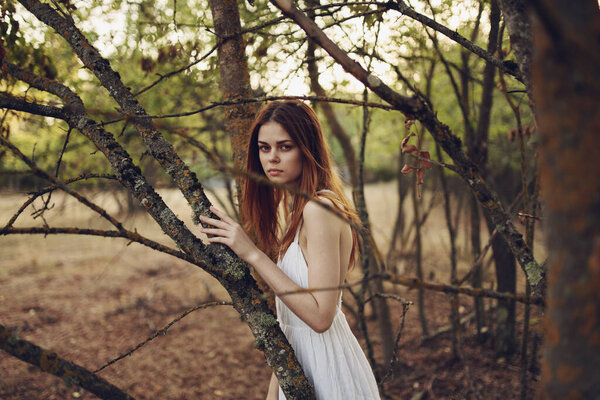 Happy red-haired woman in white dress runs on dry grass in nature near trees. High quality photo