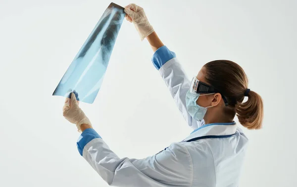 En kvinnlig läkare i medicinsk mask undersöker en röntgen på en ljus bakgrund — Stockfoto