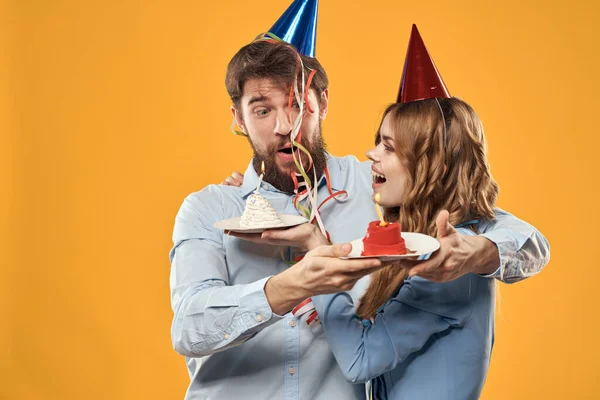 대담 한 남자와 예쁜 여자 생일 케이크를 들고 노란색 배경 디스코 파티에서 모자를 쓰고 있는 모습 — 스톡 사진