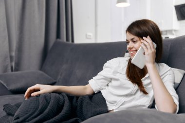 Kanepede mutlu bir kadın telefonda ve gri ekose perdelerle konuşuyor.