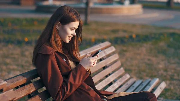 Kvinna i parken sitter på en bänk med en telefon i händerna — Stockfoto