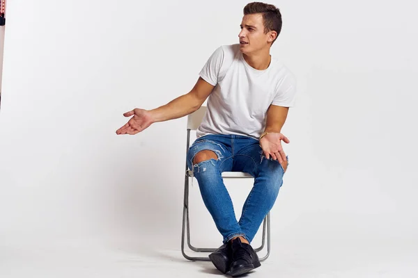 Mann auf einem Stuhl drinnen zerrissene Jeans weißes T-Shirt hübsches Gesicht Modell hellen Hintergrund — Stockfoto