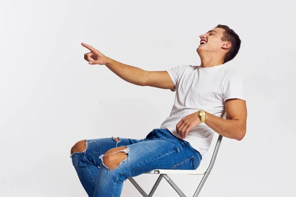 Muž na židli uvnitř roztržené džíny bílá tričko hezký obličej model světlo pozadí — Stock fotografie