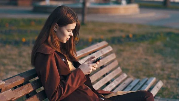Kvinna i parken sitter på en bänk med en telefon i händerna — Stockfoto