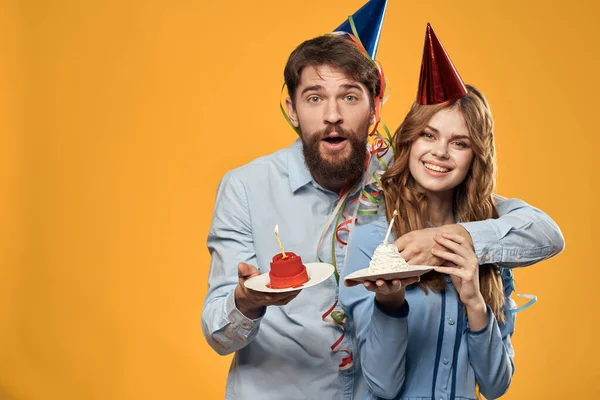 Uomo energico e bella donna con torta di compleanno in cappelli su sfondo giallo discoteca partito — Foto Stock