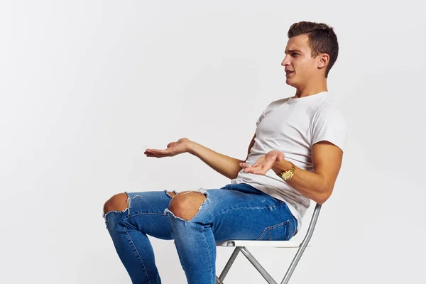 Mężczyzna na krześle wewnątrz rozdarte dżinsy biały t-shirt przystojny twarz model lekkie tło — Zdjęcie stockowe