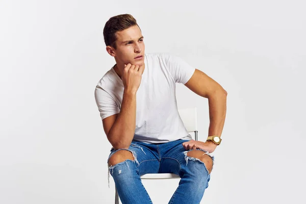 椅子の上の男室内で引き裂かれたジーンズホワイトTシャツハンサムな顔モデルライトバックグランド — ストック写真