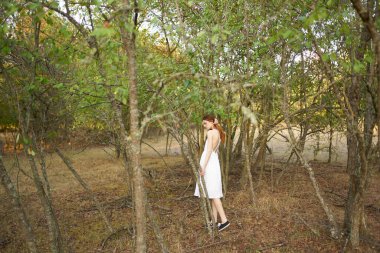 Beyaz elbiseli mutlu kızıl saçlı kadın ağaçların yanındaki kuru çimlerde koşuyor.