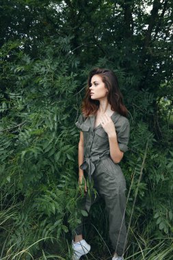 Ormanda, çalılıkların yanında yeşil tulumlu bir kadın kendi elleriyle kendine dokunuyor. 