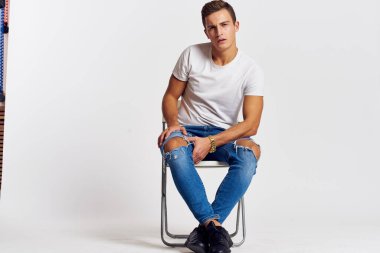 Sandalyede oturan adam yırtık kot pantolonlu, beyaz tişörtlü yakışıklı, açık renk arka planlı.