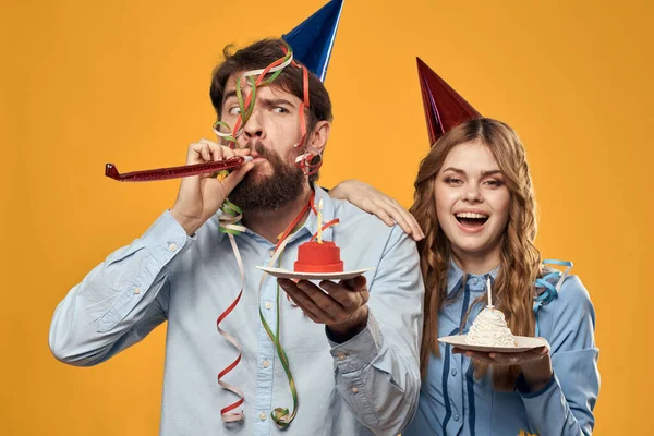 Homem e mulher em uma festa em bonés e com bolo de ouropel divertido fundo amarelo — Fotografia de Stock