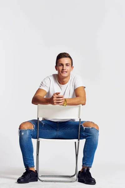 A man in jeans sits on a chair backwards and and ripped jeans T-shirt front view — Stock Photo, Image