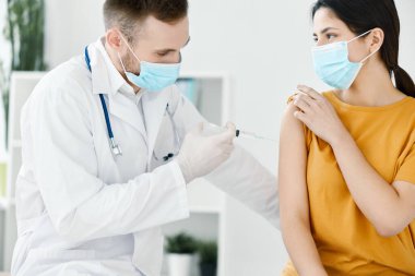 Doktor asistanı, tıbbi maske takan bir kadının omzuna iğne yapıyor.