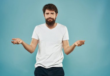 Beyaz tişörtlü ve mavi arka planda kalın sakallı bir adam elleriyle el kol hareketleri yapıyor.