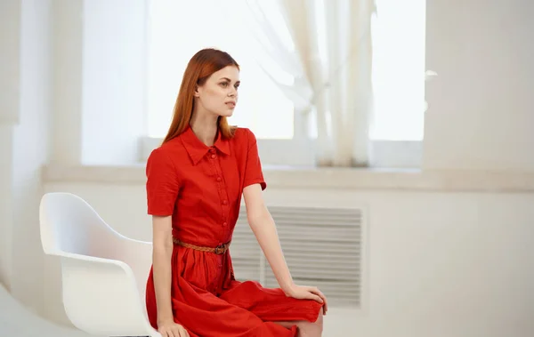 Uma mulher em um vestido vermelho senta-se em uma cadeira dentro de casa perto da janela — Fotografia de Stock