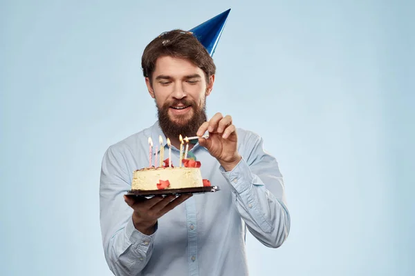 Baard man met een bord cake op een blauwe achtergrond en een verjaardagsfeestmuts op zijn hoofd — Stockfoto