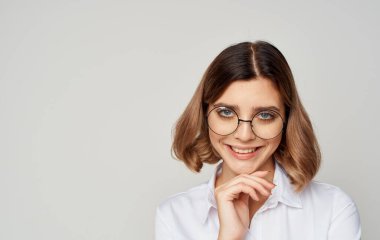 Gözlüklü, kısa saçlı, beyaz tişörtlü, bej arka planda güzel bir kadın.