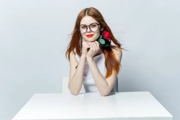 Jolie femme rousse assise à la table avec fleur de rose dans les mains glamour rouge lèvres modèle — Photo