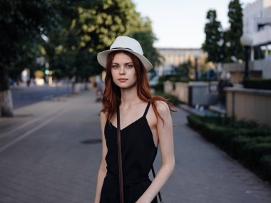 Siyah elbiseli, beyaz şapkalı, şık bir kadın parkta yürüyor.