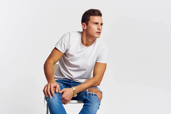 Man op een stoel binnen gescheurde jeans wit t-shirt knappe gezicht model lichte achtergrond — Stockfoto