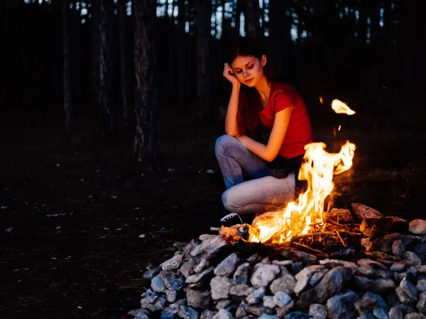 Una mujer se sienta cerca del fuego y mira el fuego en un bosque de pinos —  Fotos de Stock