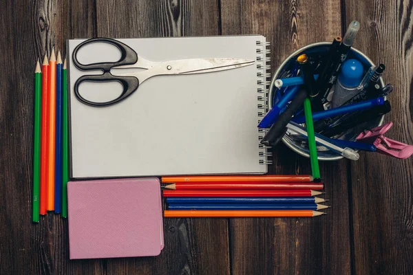 Scissors lie on an open notebook and colored pencils on a wooden table top view — Stock Photo, Image