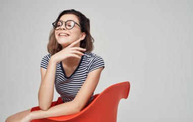 Etekli ve çizgili tişörtlü bir kadın parlak bir odada kırmızı bir sandalyede oturuyor.