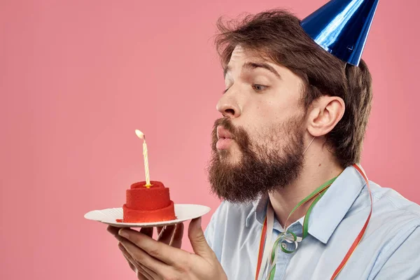 Hombre con un pastel con una vela y una gorra de cumpleaños sobre un fondo rosa — Foto de Stock