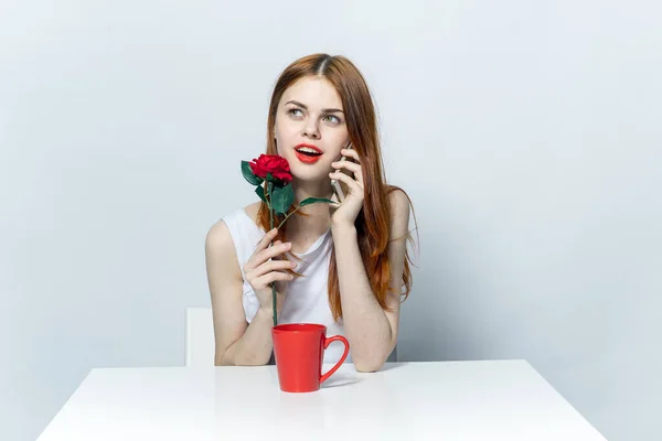 Vacker kvinna sitter vid bordet med röd ros blomma och talar i telefon — Stockfoto