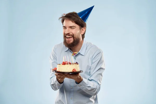 Hombre barbudo con un plato de pastel sobre un fondo azul y un sombrero de fiesta de cumpleaños en su cabeza — Foto de Stock