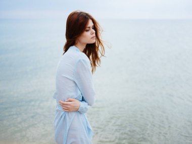 Okyanus kıyısındaki kumsalda mavi elbiseli, kızıl saçlı bir kadın.