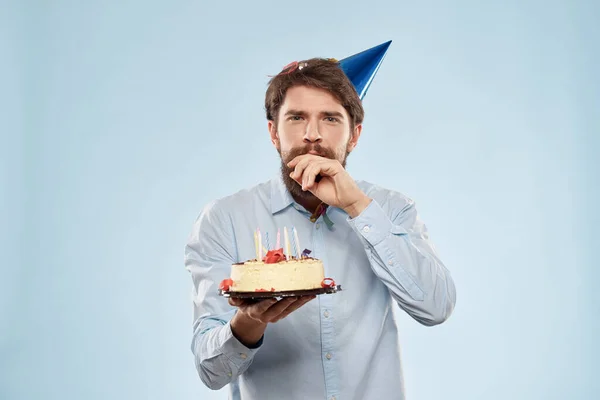 Hombre barbudo con un plato de pastel sobre un fondo azul y un sombrero de fiesta de cumpleaños en su cabeza — Foto de Stock