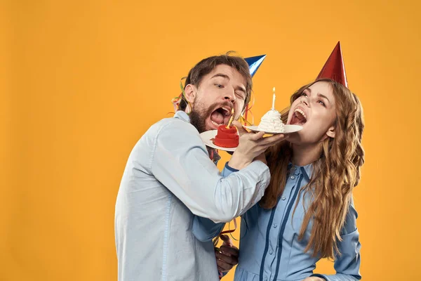 Hombre enérgico y mujer bonita con pastel de cumpleaños en sombreros en una fiesta disco de fondo amarillo — Foto de Stock
