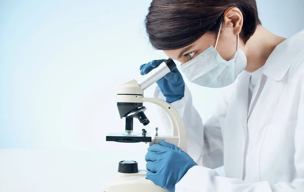 Doctora con una bata médica y guantes mirando a través de un microscopio en una mesa en el interior — Foto de Stock