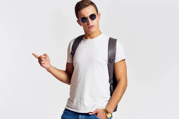 Man in zonnebril met een rugzak op zijn rug en in een wit t-shirt op een lichte achtergrond bijgesneden weergave van emotie — Stockfoto