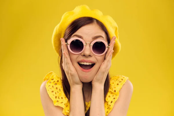 Happy woman in a yellow hat and fashionable glasses holds her hands near her face on a yellow background cropped view of emotions fun