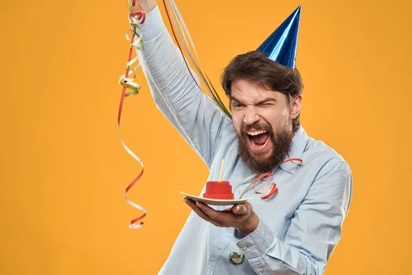 Hombre alegre con un pastel en un fondo amarillo cumpleaños días festivos gorra en su cabeza —  Fotos de Stock