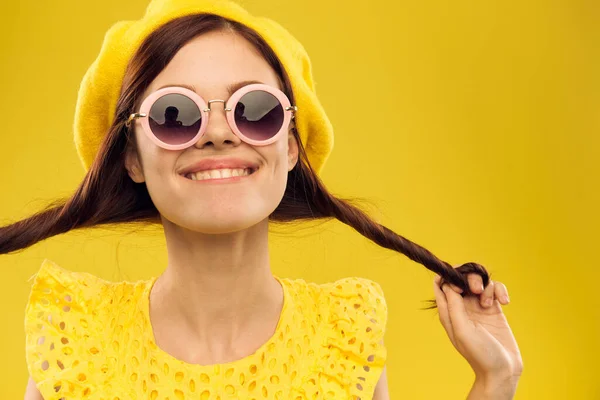 Happy woman in a yellow hat and fashionable glasses holds her hands near her face on a yellow background cropped view of emotions fun